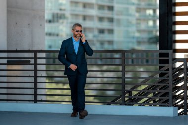 İş adamı telefonda konuşuyor ve yürüyor. Kendine güvenen bir iş adamı dışarıda telefonla konuşuyor. İş telefonu konuşması. Şirket lideri telefonda konuşmakla meşgul. Zengin iş adamı. İş çağrısı