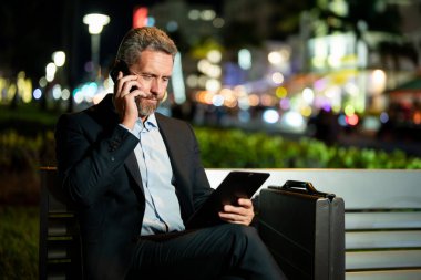 Takım elbiseli bir adam gece telefonda konuşuyor. Gece sokağında bir iş adamı arama yapıyor. Tabletli ve telefonlu bir iş adamı. Gece sokağında bankta oturan bir iş adamı. Gece işi.