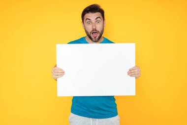 Boş afişi olan yakışıklı bir adam stüdyo arka planında izole edilmiş. Boş poster tutan şaşırmış bir adamın portresi. Boş afiş, reklam panosu ya da afiş gösteren hayret verici bir adam.