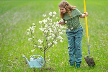 Çocuk dışarıya bir bahar ağacı dikiyor. Çocuklar meyve ağacı gibi büyür. Kürekli çocuk bahçıvan. Çocuk meyve bahçesinde çiçek açan ağaçlarla çalışıyor. Çocuklar arka bahçeye ağaç dikiyorlar. Ağaçlarla ilgilenen genç çiftçi