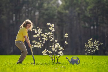 Bahçeye ağaç dikmiş bir çocuk. Çocuk kürekle toprak kazıyor ve ağaç dikiyor. Çocuk bahçıvan meyve ağacıyla ilgileniyor. Çocuklarla ağaç dikmek. Yeşillik için ağaç diken çocuk. Çocuklar için bahçe.