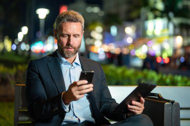 Takım elbiseli bir adam gece telefonda konuşuyor. Gece sokağında bir iş adamı arama yapıyor. Tabletli ve telefonlu bir iş adamı. Gece sokağında bankta oturan bir iş adamı. Gece işi.