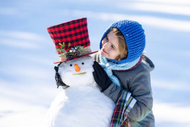 Kardan adam suratlı çocuk. Şapka ve eşarp takan çocuk. Mutlu çocuk kış günü dışarıda eğleniyor. Kardan adam yapan çocuk. Dışarıda kardan adam yapan çocuk. Çocuk yüzü