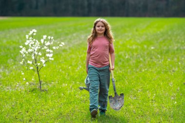 Çocuk bahçıvan arka bahçede ağaç dikiyor. Bitkilerin bahar etkinliği. Çocuk kürek ve su bidonuyla çalışıyor. Çocuk meyve ağacı dikmiş. Bahçıvan çocuklar. Çocuk yetiştirme bitkisi ve ağaç bakımı