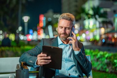 Takım elbiseli bir adam telefonda konuşuyor ve tablet kullanıyor. Tabletli bir iş adamı. Tabletli ve telefonlu bir iş adamı. Tablet kullanan bir işadamı, bankta oturur. Gece işi.