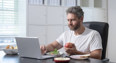 Gündelik çalışanlar masaüstünde çalışırken besleyici bir yemeğin keyfini çıkarırlar. Ev işi konsepti. Adam kahvaltı yapıyor ve uzaktan çalışıyor. Serbest çalışan biri dizüstü bilgisayarında yazarken meyve ve tahıl yer.