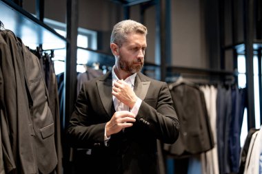 Olgun bir adam kıyafeti için mükemmel bir takım seçer. İyi giyimli bir erkek model bir butikte poz veriyor. Perakende satış asistanı şık bir adamın mükemmel takımı bulmasına yardım eder. Mağaza müdürü şık bir takım elbise ayarlar.