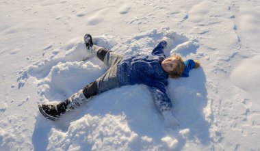 Kışlık giysili çocuk kış tatilinde kar yığınında yatıyor ve gülüyor. Kış çocukları mutluluğu. Karda yatan tatlı, heyecanlı çocuk. Çocuk dışarıda kar yağışıyla oynuyor. Çocuklar için kış aktiviteleri