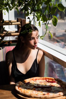Restoranda pizzayla pencerenin kenarında oturan genç bir kadın. Beyaz kadın portresi, üzerinde pizza ve gölgeler var. Siyah elbiseli bir kadın öğle yemeği için kafede pizza yiyor. Modern iç mekanda kadın ve pizza.