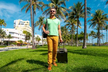 Miami sahilinde bavulla yürüyen bir turist. Yaz tatilinde valizli bir turist. Tropikal şehirde bir turist. Tropik tatil beldesinde bavullu bir turist. Turizm ve seyahat kavramı