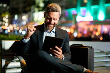 Açık hava işi. Dışarıda tablet üzerinde çalışan bir iş adamı. Tabletli bir iş adamı. Gece sokağında tabletli bir iş adamı. Tablet kullanan bir işadamı, bankta oturur. İnternette gece işi