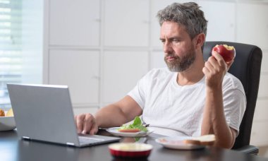 Yetişkin bir adam sabahları laptopuyla çalışırken evde kahvaltı yapar. Serbest çalışan biri, modern bir masada kahveli sıcak bir yemeğin tadını çıkarır. Çalışan öğle yemeği yerken uzaktan çalışıyor.