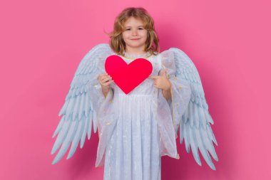 Çocuk meleğin işaret ettiği kırmızı kalp, işaret işareti. Şirin melek çocuk, stüdyo resmi. Sarışın, kıvırcık, melek kanatlı, soyutlanmış bir çocuk.