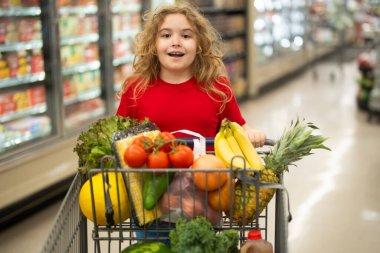 Süpermarkette el arabasıyla çocuk alışverişi. Çocuk markette taze sebze seçiyor. Yiyecek satan bir çocuk. Markette ürün seçen bir çocuk. Bakkalda taze yiyeceği olan çocuk