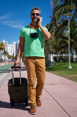 Miami sokaklarında kamera ve bavul taşıyan bir gezgin. Bir erkek gezgin, tropik bir şehirde yaz tatilinin tadını çıkarır. Yaz şehrinde bavullu bir turist. İnsan gezgin şehirde yürür