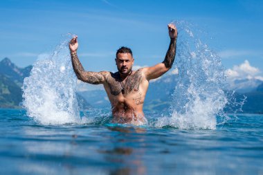 Cool water splashes around. Tattooed male stays refreshed. Powerful splash surrounds man. Sunlight hits tattoos. Summer freedom outdoors. Action moment in blue water. Athletic male expression
