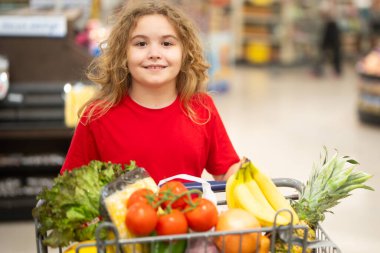 Süpermarkette el arabasıyla çocuk alışverişi. Çocuk markette taze sebze seçiyor. Süpermarkette yiyeceği olan bir çocuk. Süpermarkette ürün seçen bir çocuk. Süpermarkette taze yiyeceği olan çocuk