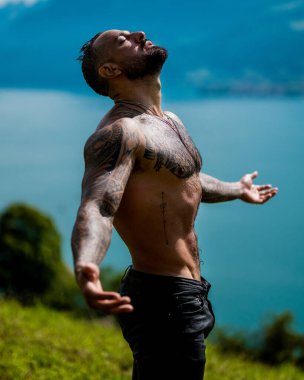 Kaslı adam temiz hava soluyor. Ellerini kaldırıp hava soluyan yakışıklı bir üstsüz model. Seksi adam doğa üzerine meditasyon yapıyor. Şelalenin yanında sakin bir nefes al. Doğada sağlık ve nefes alma.