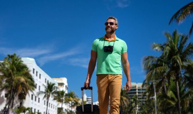 Miami sokaklarında bavulu ve kamerası olan bir turist. Bir erkek turist, tropikal bir şehirde yaz tatilinin tadını çıkarır. Yaz şehrinde bavullu bir turist. Erkek turist şehirde yürüyor