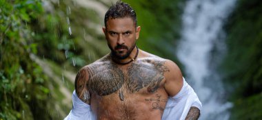 Strong man standing under waterfall rain. Confident man with tattooed body. Intense man portrait with water drops. Powerful man expression in nature