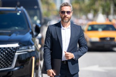 Dışarıdaki iş adamı. Zengin iş adamı başarının tadını çıkarıyor. Şık takım elbiseli lüks iş adamı. Pahalı arabanın önündeki zengin adam. Lüks iş seyahati. Klasik takım elbiseli, kendinden emin zengin bir iş adamı.