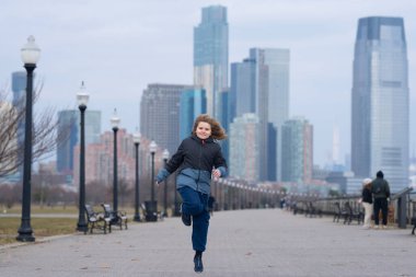 Nehir kenarında koşan bir çocuk. Çocuk dışarıda sağlıklı bir egzersizin parçası olarak koşuyor. Kamusal alanda enerji ve neşe içinde koşan bir çocuk. Modern bir şehir ortamında koşan çocuk