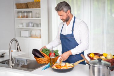 Mutfakta ev yapımı yemek için malzeme doğrayan bir adam. Bir şef mutfakta vejetaryen yemeği hazırlıyor. Çiğ sebzeli bir aşçı. Önlüklü bir adam evde besleyici bir öğle yemeği için sebze doğruyor.
