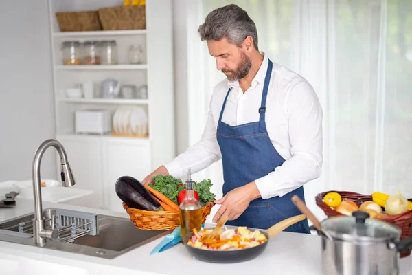 Mutfakta ev yapımı yemek için malzeme doğrayan bir adam. Bir şef mutfakta vejetaryen yemeği hazırlıyor. Çiğ sebzeli bir aşçı. Önlüklü bir adam evde besleyici bir öğle yemeği için sebze doğruyor.