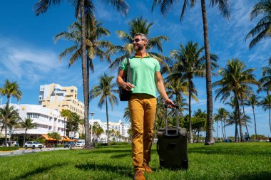 Miami sahilinde bavulla yürüyen bir turist. Yaz tatilinde valizli bir turist. Tropikal şehirde bir turist. Tropik tatil beldesinde bavullu bir turist. Turizm ve seyahat kavramı