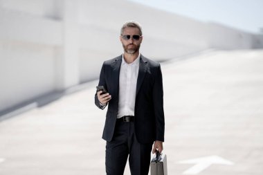 İş portresi. Dışarıdaki telefondan iş adamı aradı. Başarılı lider hareket halinde. Takım elbiseli, çantalı bir adam telefonda konuşuyor. İşadamı işe gidiyor. Çantası olan bir iş adamı. İş başarısı