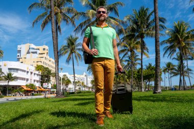 Miami sahilinde bavulla yürüyen bir turist. Yaz tatilinde valizli bir turist. Tropikal şehirde bir turist. Tropik tatil beldesinde bavullu bir turist. Turizm ve seyahat kavramı