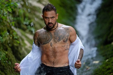 Şelale yağmurunun altında duran güçlü adam. Dövmeli, kendinden emin bir adam. Su damlaları ile yoğun bir adam portresi. Doğadaki güçlü insan ifadesi.