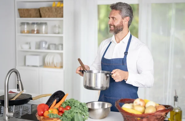 Taze sebze pişiren bir şef. Ev yapımı yemek pişiren bir adam. Mutfakta sağlıklı bir kahvaltı için sebze pişiren bir aşçı. Üniformalı bir adam beyaz bir evde sebze pişiriyor.