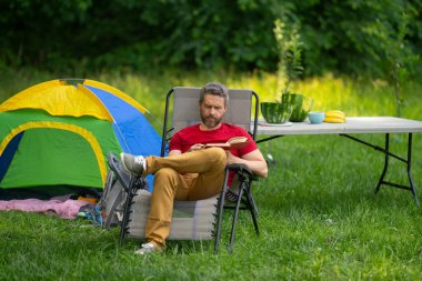 Gezgin doğada kamp yapmaktan hoşlanıyor. Doğaya yaz gezisi. Orman kampında dinlenen adam. Kamp gezisi. Yürüyüşten sonra çadırın yanında dinleniyor. Barışçıl bir kamp alanında özgürlük tatili. Yazın piknikteki adam