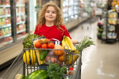 Süpermarkette el arabasıyla çocuk alışverişi. Çocuk markette taze sebze seçiyor. Yiyecek satan bir çocuk. Markette ürün seçen bir çocuk. Bakkalda taze yiyeceği olan çocuk