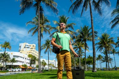 Miami sahilinde bavulla yürüyen bir turist. Yaz tatilinde valizli bir turist. Tropikal şehirde bir turist. Tropik tatil beldesinde bavullu bir turist. Turizm ve seyahat kavramı