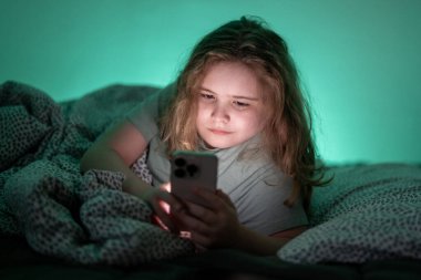 Çocuk gece yatakta dijital telefon kullanıyor. Çocuk geceleri telefonda sosyal medyayı karıştırıyor. Ergen gece yatakta telefonla dinleniyor. Çocuk gece yatakta telefonla emilmiş.