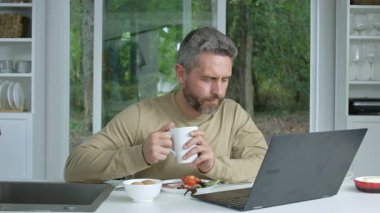 Sabah mutfağı, kahvaltı ve dizüstü bilgisayarı açık bir adamla dolu. Adam mutfakta oturup dizüstü bilgisayar kullanırken yemek yiyor. Laptop işi ve kahvaltı rahat mutfağın içinde yeniyor.
