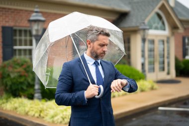 İş adamı elinde şemsiyeyle yağmurun altında bekliyor. Kendine güvenen bir adam ıslak bir sokakta dururken saatini kontrol eder. İş adamı saati kontrol ediyor. Saati izleyen adam. Dakik bir iş adamı.