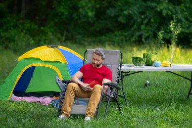 Kampta kitap okuyan bir adam. Doğaya yaz gezisi. Orman kampında dinlenen adam. Kamp gezisi. Yürüyüşten sonra çadırın yanında dinleniyor. Barışçıl bir kamp alanında özgürlük tatili. Piknikteki Adam