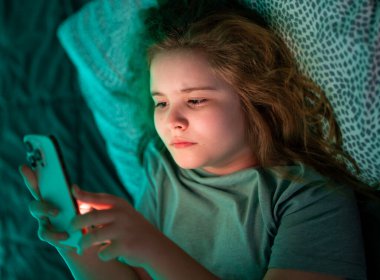 Çocuk gece yatağında telefon kullanıyor. Geceleri elinde telefon tutan bir çocuk. Ergen telefonları geceleri yatakta kaydırıyor. Çocuk gece yatakta telefon ekranını izliyor.