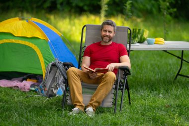 Kampta kitap okuyan bir adam. Doğaya yaz gezisi. Orman kampında dinlenen adam. Kamp gezisi. Yürüyüşten sonra çadırın yanında dinleniyor. Barışçıl bir kamp alanında özgürlük tatili. Piknikteki Adam