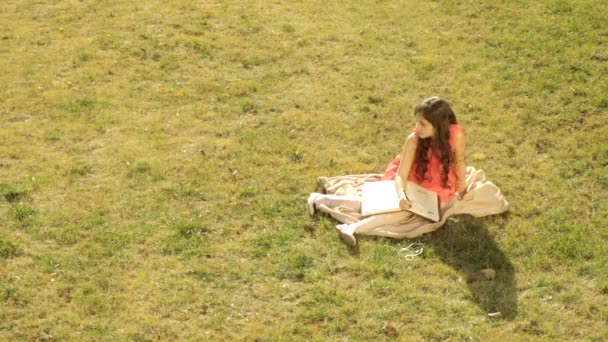 Liten flicka sitter med en anteckningsbok på en matta i trädgården — Stockvideo