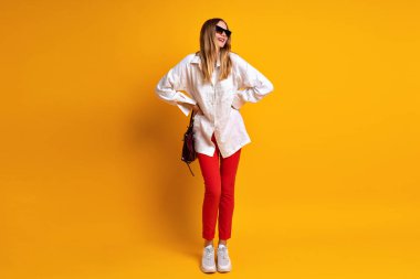 Kırmızı pantolon ve beyaz gömlek giyen şık bir iş kadını, şık çantalı güneş gözlüğü ve aksesuar takan bir stüdyo resmi. sarı arkaplan, boşluğu kopyala.