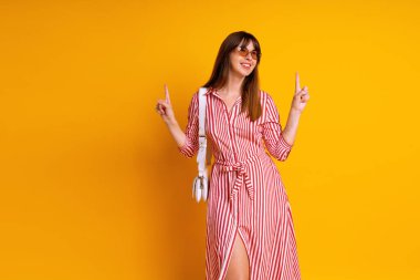 Çizgili kırmızı elbise, bej güneş gözlüğü ve beyaz çanta giyen şık bir kadının kapalı mekan portresi, stüdyo sarısı..  