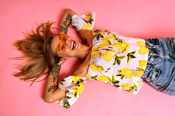 ポジティブな夏のスタジオポートレートの陽気ブロンド女性在床の上に身に着けているトレンディーなサングラス小さく笑って トレンディーなブロガーの衣装 ピンクの背景 — ストック写真