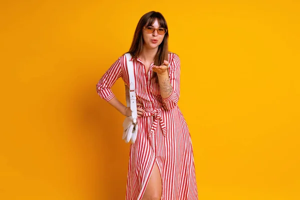 穿着条纹红色连衣裙 米色太阳镜和白色包袋 时尚气质 工作室黄色背景的时髦女人的室内肖像 — 图库照片