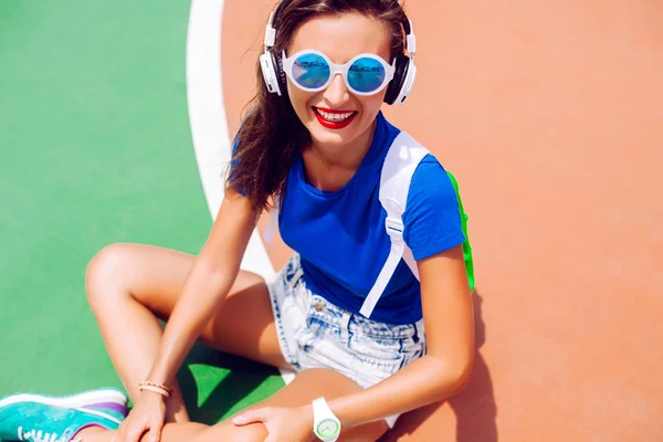 Levensstijl heldere mode portret van jonge fit stijlvolle vrouw die zich voordeed op grond van de sport, het dragen van trendy neon swag look, zonnebril en rugzak, luisteren haar favoriete muziek op grote oortelefoons — Stockfoto