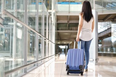 Beyaz gömlek ve kotlu Asyalı bir kadın yolcunun arka görüntüsü, mavi bir bavulu yuvarlıyor, havaalanındaki terminaline doğru yürüyor, yan tarafında cam pencereler var..