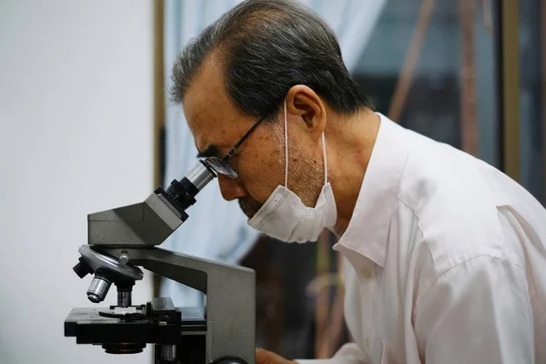 A old Asian doctor with glasses and face mask is using his microscope to inspect his patient blood sample, analyzing the red blood cell for any abnormality.